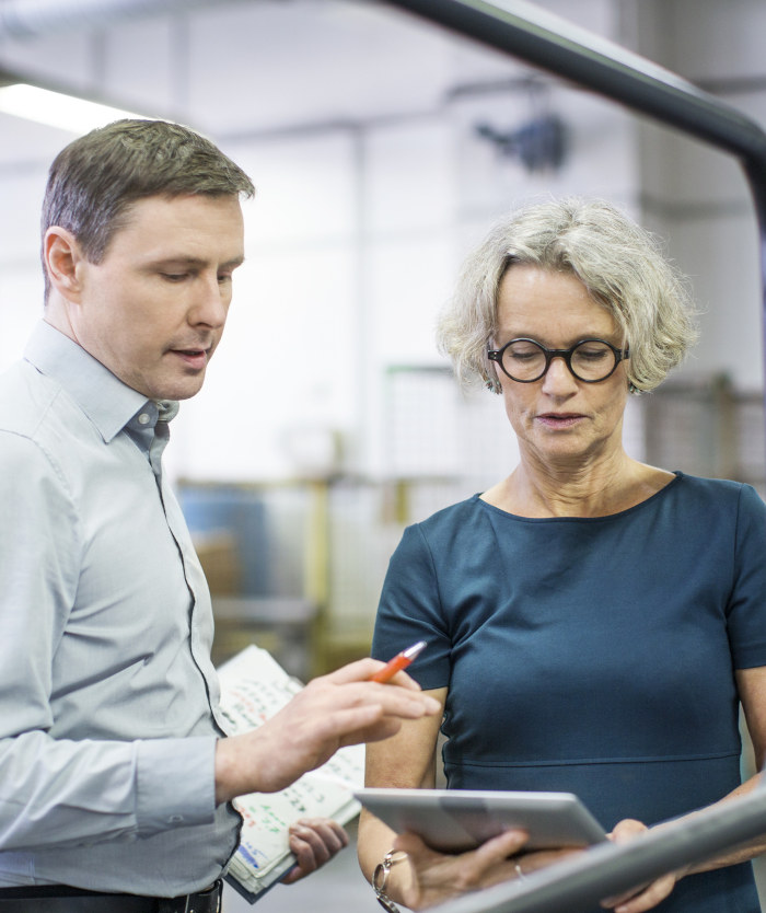 Deux personnes en discussion dans un environnement professionnel, tenant des documents et une tablette, illustrant la mise en place des QCE.